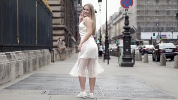 Boldog fiatal nő fehér ruhában pózol a háttérben város utcáin. Akció. Vonzó szőke mosolygós és pózol a fehér ruha ellen utcában fények és múló autók — Stock videók