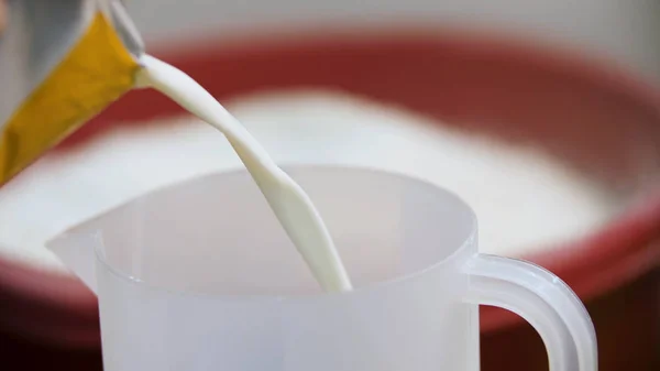 Derramando líquido branco para um copo de medição com um recipiente de farinha no fundo, conceito de cozinha. Imagens de stock. Leite sendo derramado em copo de medição de plástico . — Fotografia de Stock