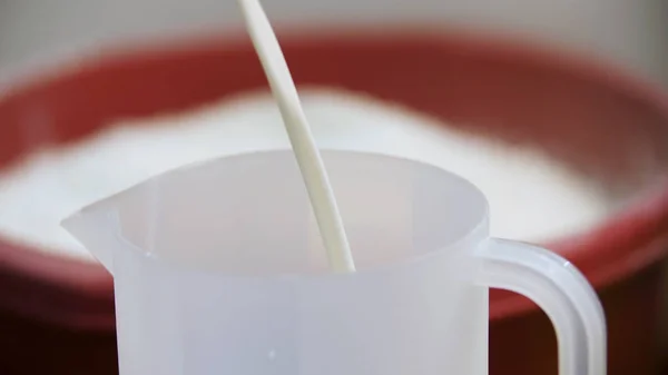흰색 액체를 계량 컵에 붓고 밀가루 용기와 함께 배경에 요리 개념. 스톡 푸티지. 플라스틱 측정 컵에 붓는 우유. — 스톡 사진