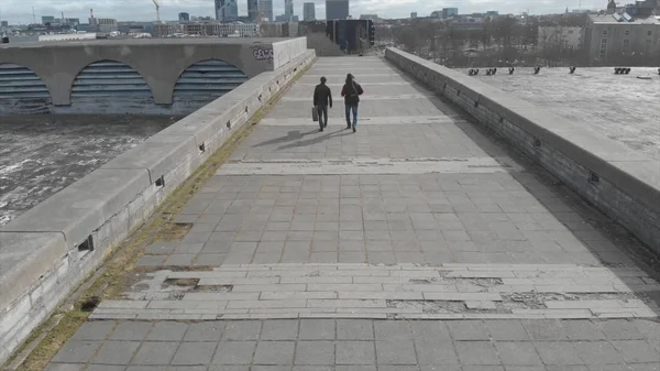 Vista trasera de dos hombres con guitarras caminando sobre un gran puente gris contra el cielo azul nublado y edificios de la ciudad a principios de primavera. Acciones. Arquitectura urbana — Foto de Stock