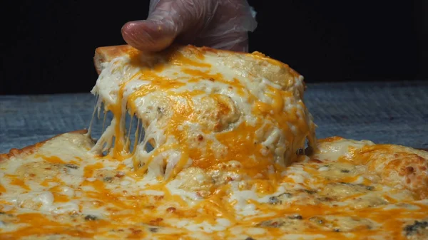 Крупный план руки шеф-повара в защитной перчатке, который берет вкусный кусок пиццы с 4 сырами, лежащий на деревянной доске. Рамка. Традиционная итальянская кухня — стоковое фото