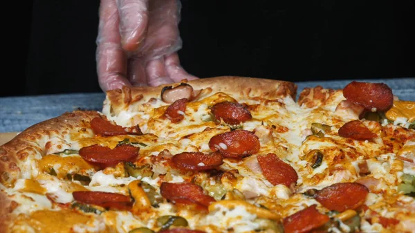 Крупный план руки шеф-повара в защитной перчатке, принимающего вкусный кусок пиццы с салями, огурцы сыра на деревянной доске. Рамка. Традиционная итальянская кухня — стоковое фото