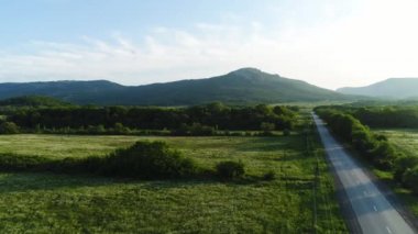 Arka planda fantastik dağlar ile yeşil çim alanları peyzaj. Atış. Yeşil çayırlar, çalılar ve parlak gökyüzü ile ağaçlar, doğanın güzelliği ile asfalt yol.