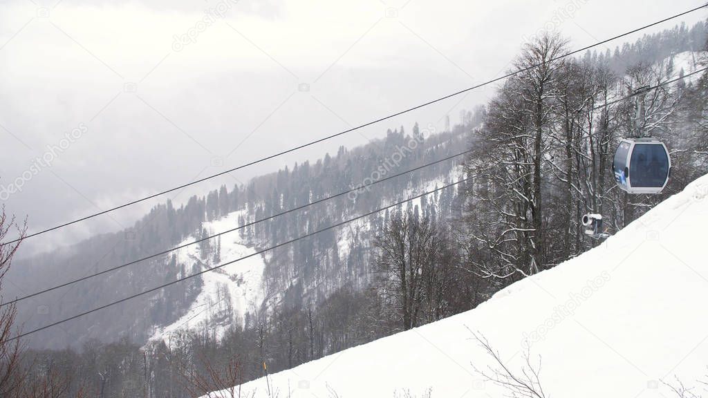 Winter landscape with mountain slope covered by snowy trees and the funicular with moving cabins, ski resort. Art. Ski cable car over the valley on grey sky background.