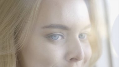 Güzel mavi gözlü genç kadının yakın çekim izliyor ve gülümsüyor. Eylem. Büyüleyici mavi gözleri ile Lovely sarışın bayan gülümsüyor ve açık pencere önünde büyüleyici görünüyor