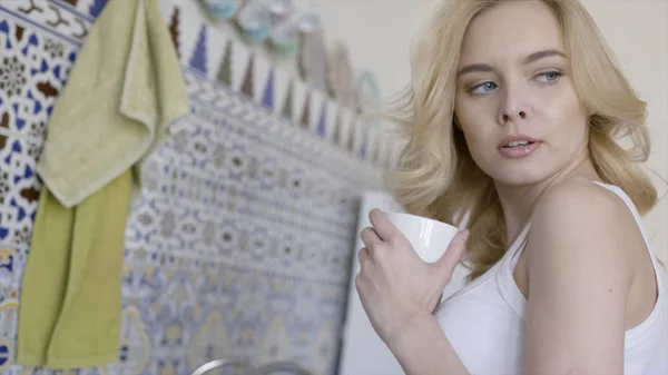 Woman takes mug on kitchen background. Action. Beautiful young blonde woman with blue eyes takes mug with hot drink and throws charming look back — 스톡 사진