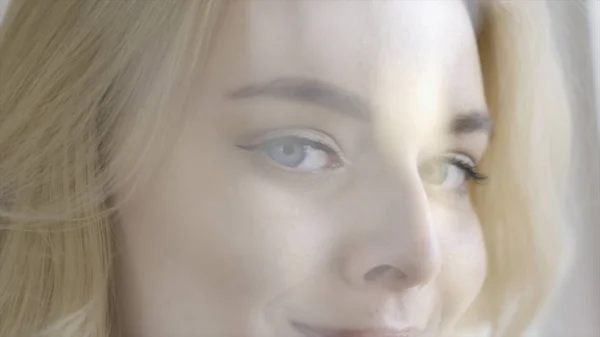 Close-up de bela mulher jovem de olhos azuis assistindo e sorrindo. Acção. Linda senhora loira com encantadores olhos azuis sorrisos e olha encantadoramente na frente da janela clara — Fotografia de Stock