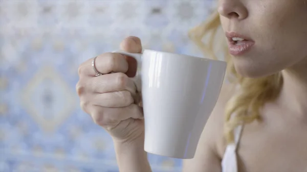 Belle jeune femme blonde aux yeux bleus qui boit du thé. L'action. Jeune belle dame avec anneau sur son doigt boire du thé chaud parfumé de tasse blanche — Photo