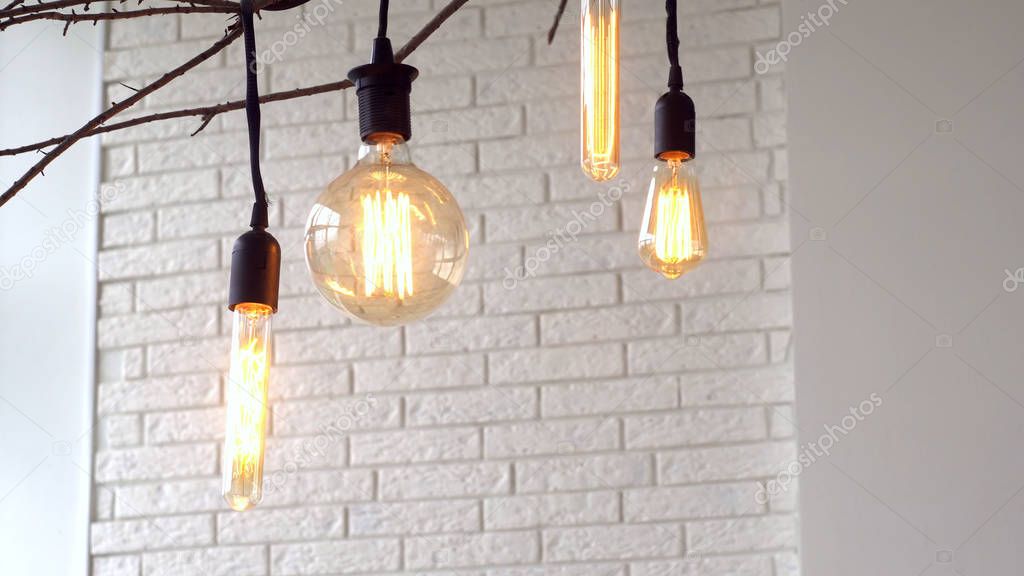 Vintage hanging lamps on white background of wall. Media. Glowing vintage light bulbs of different shapes hang on branch in interior with white brick wall