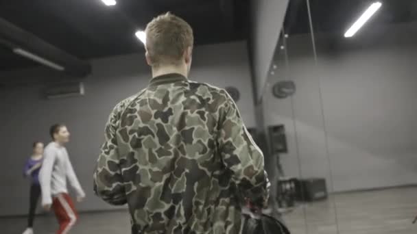 Achteraanzicht van een man in camouflage jas wandelen langs de spiegel muur in dans klasse. Actie. Jonge man komt naar Dance Hall waar andere peiple-trein. — Stockvideo