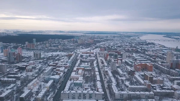 Vista aérea de los edificios de la ciudad cubiertos de nieve, carreteras y gran río en el fondo contra el cielo gris de la noche en invierno. Clip. Invierno ciudad paisaje — Foto de Stock