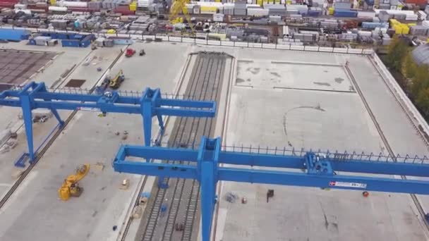 Aérien pour grue à portique géant bleue debout sur les rails et les fondations en béton. Clip. Vue de dessus de l'entrepôt avec des machines, des conteneurs et des travailleurs . — Video