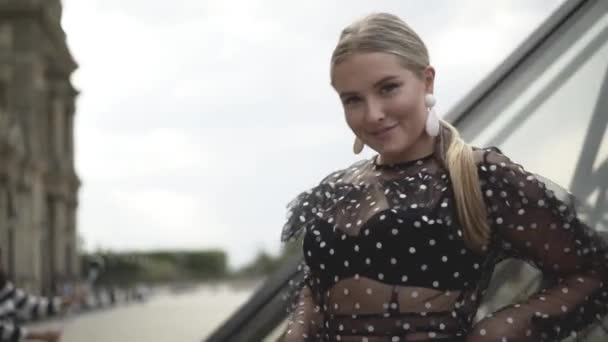 Belle jeune femme regarde la caméra avec un regard charmant sur le fond des bâtiments architecturaux. L'action. Belle blonde posant en chemisier transparent sur fond de rues de la ville — Video