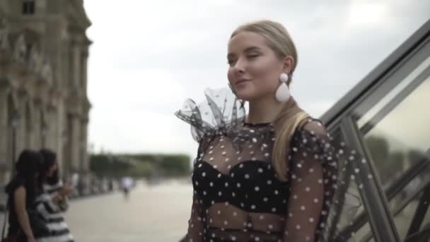 Hermosa mujer joven mira a la cámara con una mirada encantadora en el fondo de los edificios arquitectónicos. Acción. Atractiva rubia posando en blusa transparente sobre fondo de calles de la ciudad — Vídeos de Stock