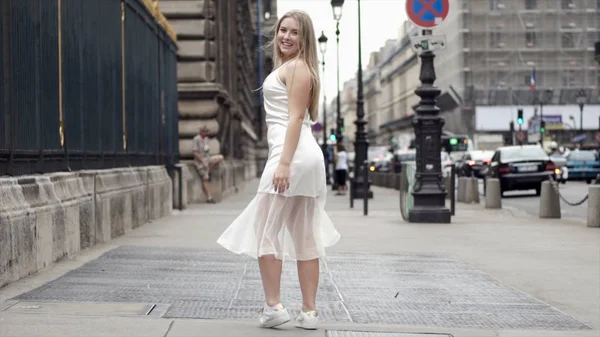 Boldog fiatal nő fehér ruhában pózol a háttérben város utcáin. Akció. Vonzó szőke mosolygós és pózol a fehér ruha ellen utcában fények és múló autók — Stock Fotó