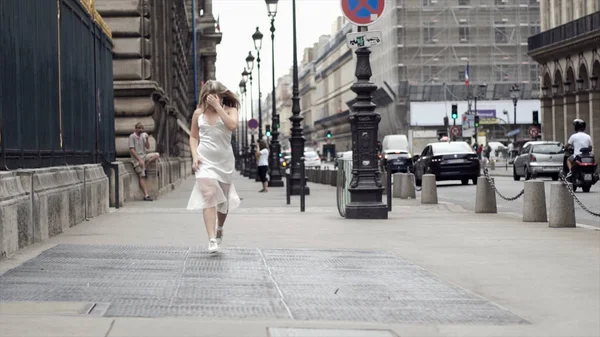 Boldog fiatal nő fehér ruhában pózol a háttérben város utcáin. Akció. Vonzó szőke mosolygós és pózol a fehér ruha ellen utcában fények és múló autók — Stock Fotó
