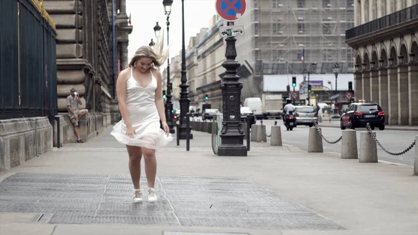 Beyaz elbiseli mutlu genç kadın arka plan şehir sokaklarında poz. Eylem. Çekici sarışın gülümseyen ve ışıklar ve geçen arabalar ile sokağa karşı beyaz elbise poz — Stok fotoğraf