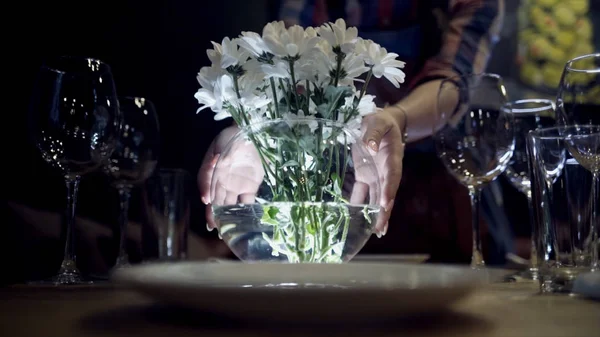 Nő hozza váza virágot asztalra. Akció. Közeli fel a nő hozza váza, fehér virágot szolgáló asztal titokzatos fények, sötét háttér — Stock Fotó