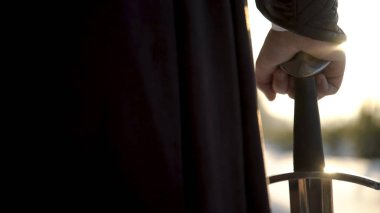 Ortaçağ takım elbiseli adam elinde kılıç. Eylem. Ortaçağ kıyafeti içinde insan el yakın çekim güneş ışığı arka planda kılıç kabza tutarak. Ortaçağ kahramanının destansı görünümü