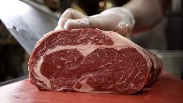 El carnicero con guantes corta la canal de la carne roja. Acción. Primer plano del cuchillo profesional cortando enorme pedazo de carne en la carnicería — Foto de Stock