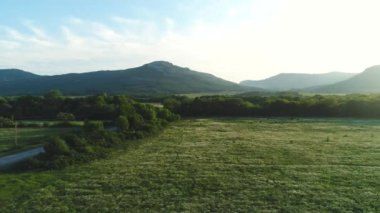 Mavi bulutlu gökyüzüne karşı yaz aylarında arka planda ağaçlar, kırsal yol ve dağlar ile güzel yeşil çayır havadan görünümü. Atış. Pitoresk yaz manzarası