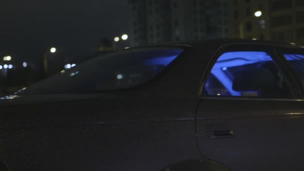 Vista lateral de un coche sintonizado con luz azul neón en el interior que se mueve en una calle de una ciudad nocturna moderna. Acción. Hermoso lila brillante vehículo de pasajeros conduciendo por la noche . — Vídeo de stock