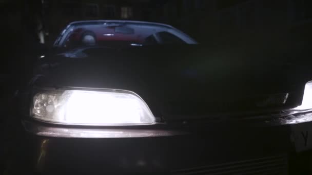 夜間に点灯する駐車中の乗用車のヘッドライト、車の詳細コンセプトをクローズアップ。アクション。車のライトをオンにするドライバーと暗闇の中に立っている車両. — ストック動画