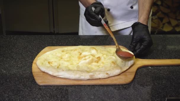 Close-up of chef prepares delicious Italian pizza in restaurant. Frame. Chef pours tomato sauce on baked round pizza base. Pizza cooked in oven on wooden tray — Stock Video