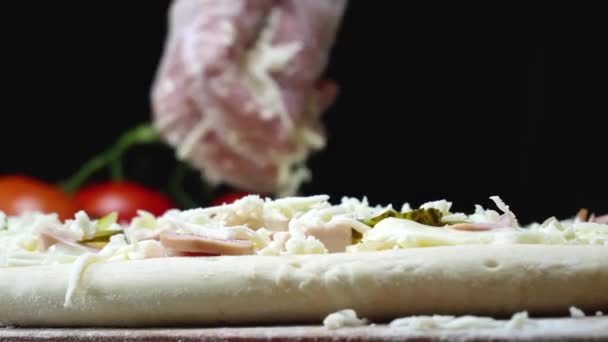 Ο σεφ ρίχνει τυρί στην πίτσα. Πλαίσιο. Ο επαγγελματίας σεφ της ιταλικής κουζίνας με γάντια ρίχνει τριμμένο τυρί σε ακατέργαστη πίτσα. Παρασκευή παραδοσιακών πίτσας σε μαύρο απομονωμένο φόντο — Αρχείο Βίντεο