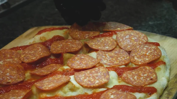 Närbild av kocken förbereder pizza pepperoni. Ram. i handskar sätter skivor av pepperoni korv på rå pizza innan stekning. Italienska matlagning pizza — Stockfoto
