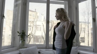 Siyah hırka kapalı alarak mavi kot pantolon ve beyaz tank üst genç ve güzel sarışın kadın yan görünümü. Eylem. Rahat daire içinde büyük pencere yakınında yürüyen kız.