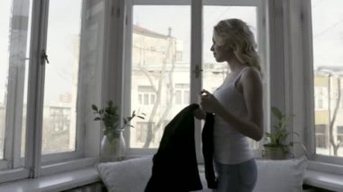 Siyah hırka kapalı alarak mavi kot pantolon ve beyaz tank üst genç ve güzel sarışın kadın yan görünümü. Eylem. Rahat daire içinde büyük pencere yakınında yürüyen kız.