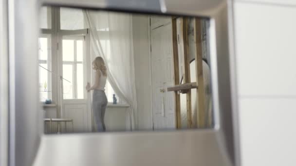 Un reflet dans le miroir d'une femme blonde mince en jeans bleus et débardeur blanc à la maison debout près de la fenêtre. L'action. Belle fille à l'intérieur de son appartement, vue arrière . — Video