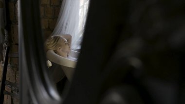 Banyo iç rahatlatıcı ve ayna yansıması saçlarını dokunan güzel bir genç sarışın kadın yan görünümü. Eylem. Pencerenin yanındaki beyaz küvette oturan bir kız..
