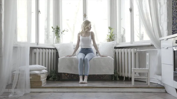 Mujer rubia bonita sentada en un pequeño sofá blanco en casa frente a la ventana. Acción. Chica atractiva en camiseta blanca y pantalones vaqueros azules en su apartamento luminoso . —  Fotos de Stock