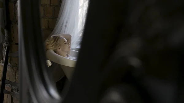 Vista lateral de una hermosa joven rubia relajándose en el interior del baño y tocando su cabello en el reflejo del espejo. Acción. Una chica sentada en la bañera blanca cerca de la ventana . — Foto de Stock