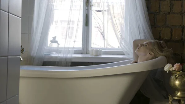 バスルームのインテリアでリラックスし、彼女の髪に触れる美しい若いブロンドの女性の側面図。アクション。窓の近くの白い浴槽に座っている女の子. — ストック写真