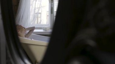 Banyo iç rahatlatıcı ve ayna yansıması saçlarını dokunan güzel bir genç sarışın kadın yan görünümü. Eylem. Pencerenin yanındaki beyaz küvette oturan bir kız..