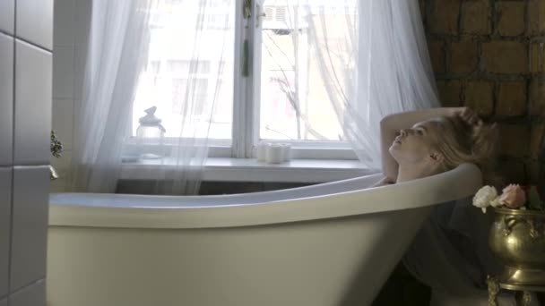 Vista lateral de una hermosa joven rubia relajándose en el interior del baño y tocando su cabello. Acción. Una chica sentada en la bañera blanca cerca de la ventana . — Vídeos de Stock