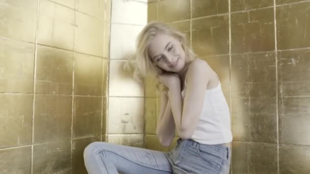 Blond model with curly hair sitting in front of golden brick wall and tilting her head. Action. Beautiful young girl smiling in the bathroom. — Stock Video