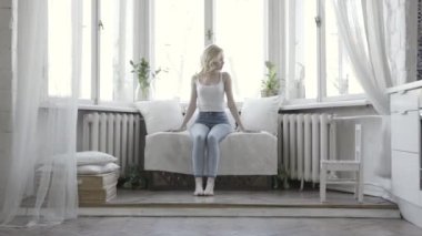 Güzel sarışın kadın pencerenin önünde evde beyaz küçük kanepede oturuyor. Eylem. Onu parlak dairede beyaz tank üst ve mavi kot çekici kız.