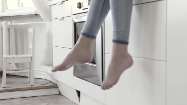 Nahaufnahme für Frauenbeine, die mit weißen Küchenschubladen und Backofen auf dem Hintergrund baumeln. Aktion. Frauen in Jeans sitzen barfuß auf dem Küchentisch. — Stockvideo