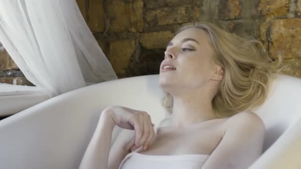 Портрет красивой блондинки в белом топе, сидящей в пустой ванне на фоне стены из красного кирпича. Начали. Молодая модель расслабляется и трогает шею и руку . — стоковое видео