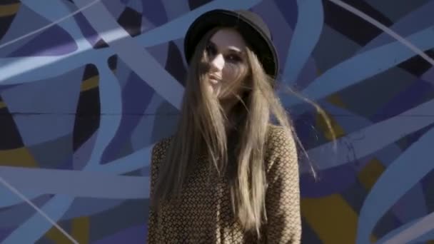 Portrait of young beautiful fashionable woman with black hat posing on blue graffity background. Action. Model wearing stylish wide-brimmed hat looking at camera, female fashion. — Stock Video