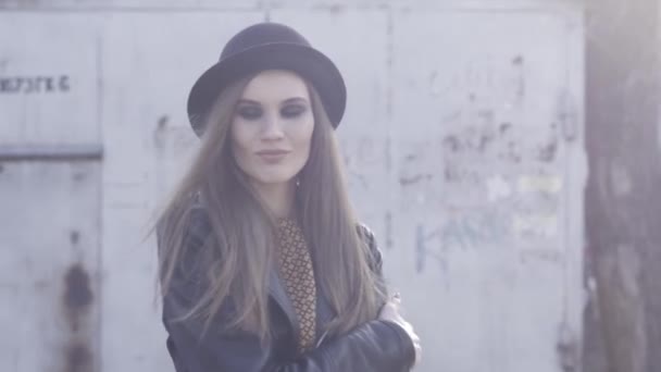 Pretty trendy girl with black stylish hat and leather jacket posing at old building in warm sunlight and hugging herself. Action. Smiling young model with smoky eyes standing outside. — Stock Video