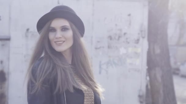 暖かい日差しの中で古い建物でポーズ黒のスタイリッシュな帽子と革のジャケットを持つかなりトレンディな女の子。アクション。スモーキーな目と外に立つ毛を持つ笑顔の若いモデル. — ストック動画