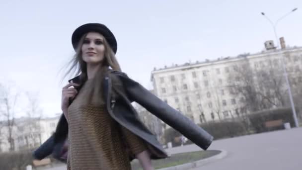 Fille marchant dans la rue avec sa veste en cuir noir sur les épaules et de longs cheveux raides flottant dans le vent. L'action. Une femme marche seule dans la ville sur fond de ciel bleu . — Video