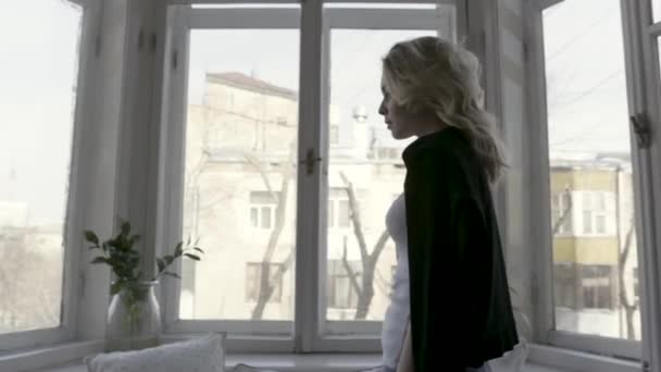 Vista laterale di una bella bionda magra a casa dalla finestra. Azione. Giovane ragazza con cardigan nero sulle spalle camminando verso la finestra . — Video Stock