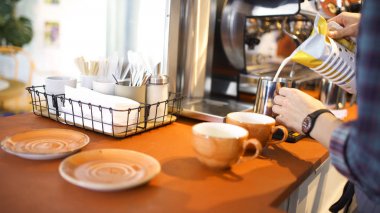 Profesyonel bir barista yan görünümü kahve dükkanında sürahi içine taze süt koyarak, sıcak içecekler hazırlık. Madde. Kahve makinesi, fincanlar, tabaklar ve kahvehanedeki diğer şeyler..