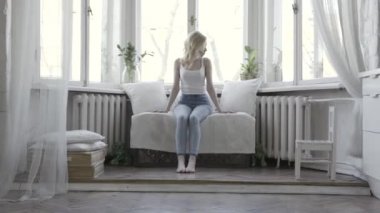 Üzgün genç kadın oturma ve onun parlak dairenin büyük pencerelerin önünde kanepede düşünme. Eylem. Evde bir kanepede mavi kot pantolon ve beyaz tank üst sarışın kadın.